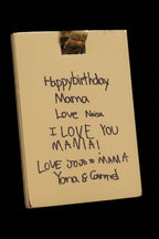 A close-up of a gold-colored rectangular card or plaque with handwritten birthday message for 'Mama' in black marker, held by a small metallic clip against a dark background.