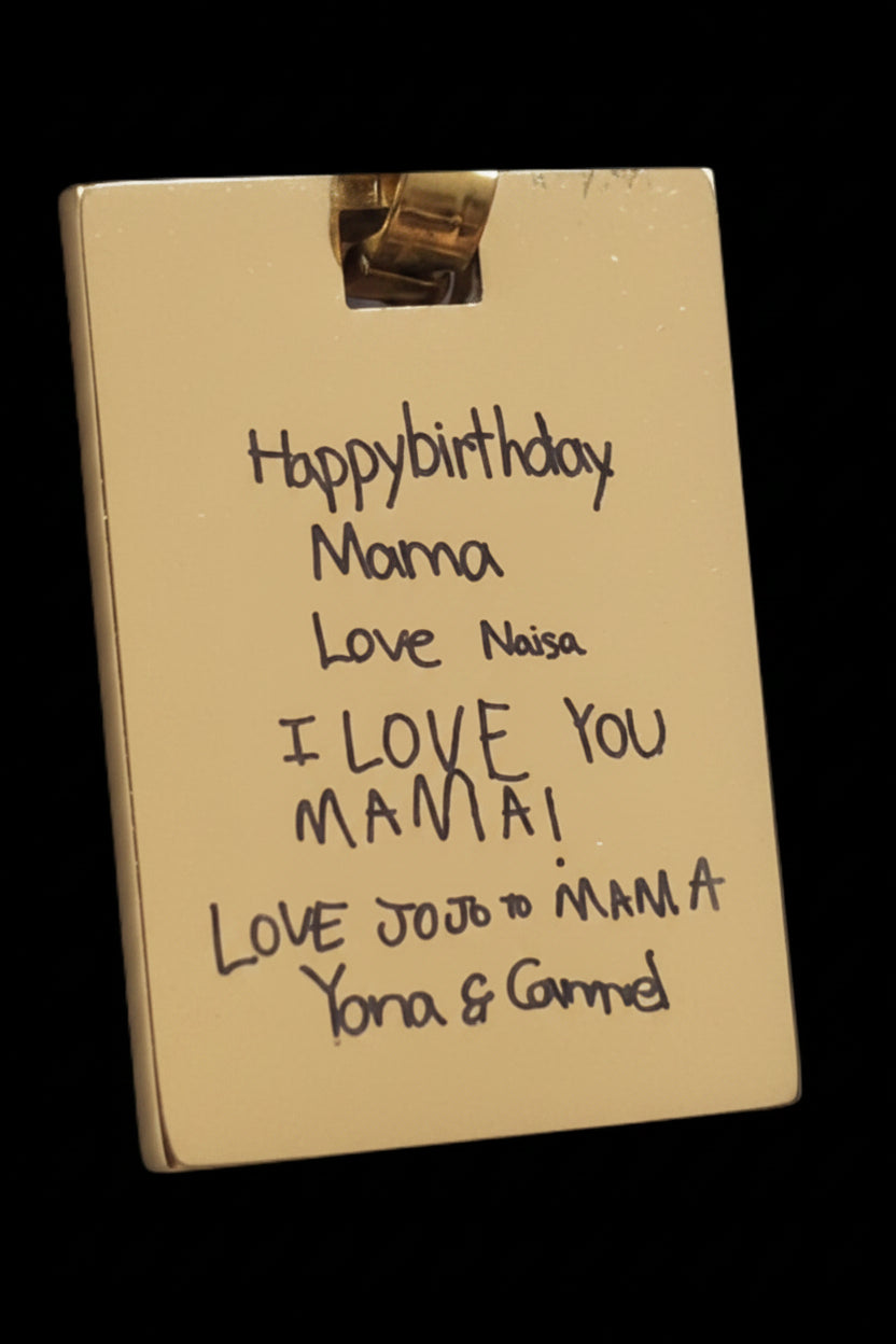 A close-up of a gold-colored rectangular card or plaque with handwritten birthday message for 'Mama' in black marker, held by a small metallic clip against a dark background.