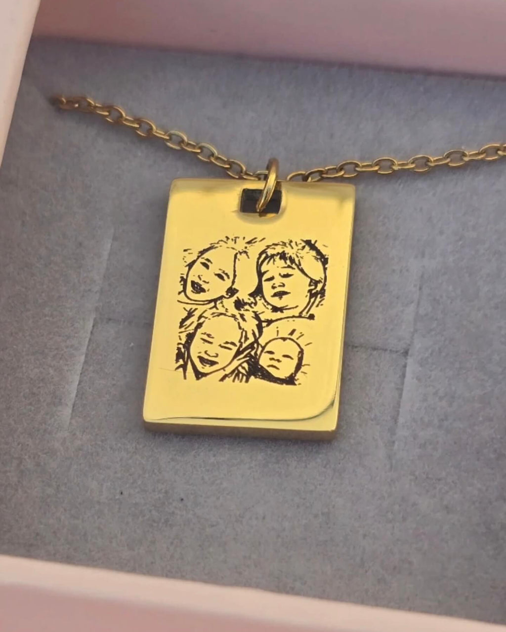 A gold rectangular pendant necklace with an engraved portrait design, resting on a gray velvet-lined pink jewelry box.