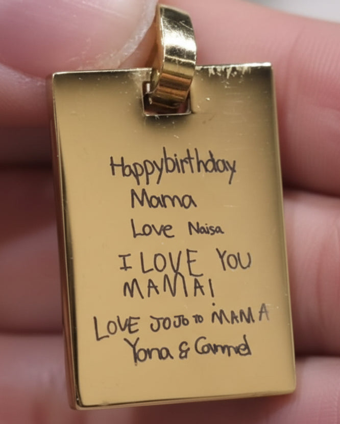 A hand holding a small engraved gold pendant or tag with a loop at the top, photographed in a real indoor setting with a blurred background.