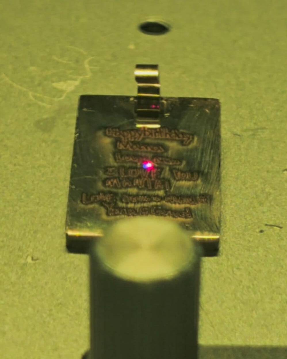 Engraving process showing a small rectangular pendant , photographed on a yellow-green surface. A bright red laser dot is visible on the metal, and part of a dark clamp or stand appears in the foreground.