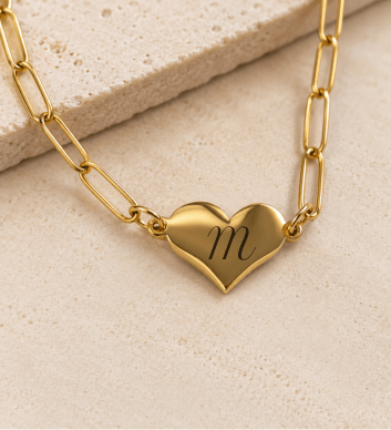 A gold paperclip chain necklace with a polished heart-shaped pendant engraved with the lowercase letter 'm', displayed on a textured beige stone surface.