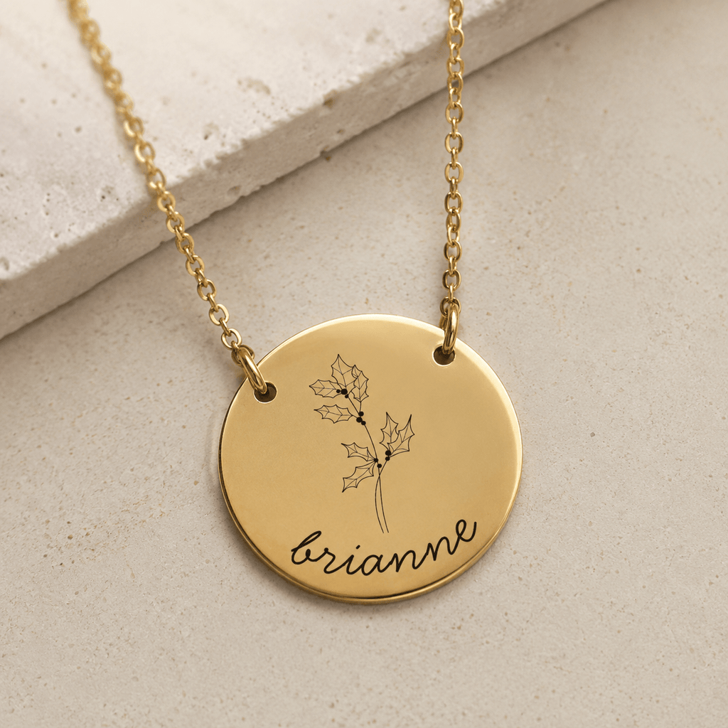 A gold round pendant necklace engraved with a holly leaf design and the name 'brianne' in script, displayed on a textured beige stone surface.