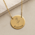 A gold round pendant necklace engraved with a holly leaf design and the name 'brianne' in script, displayed on a textured beige stone surface.