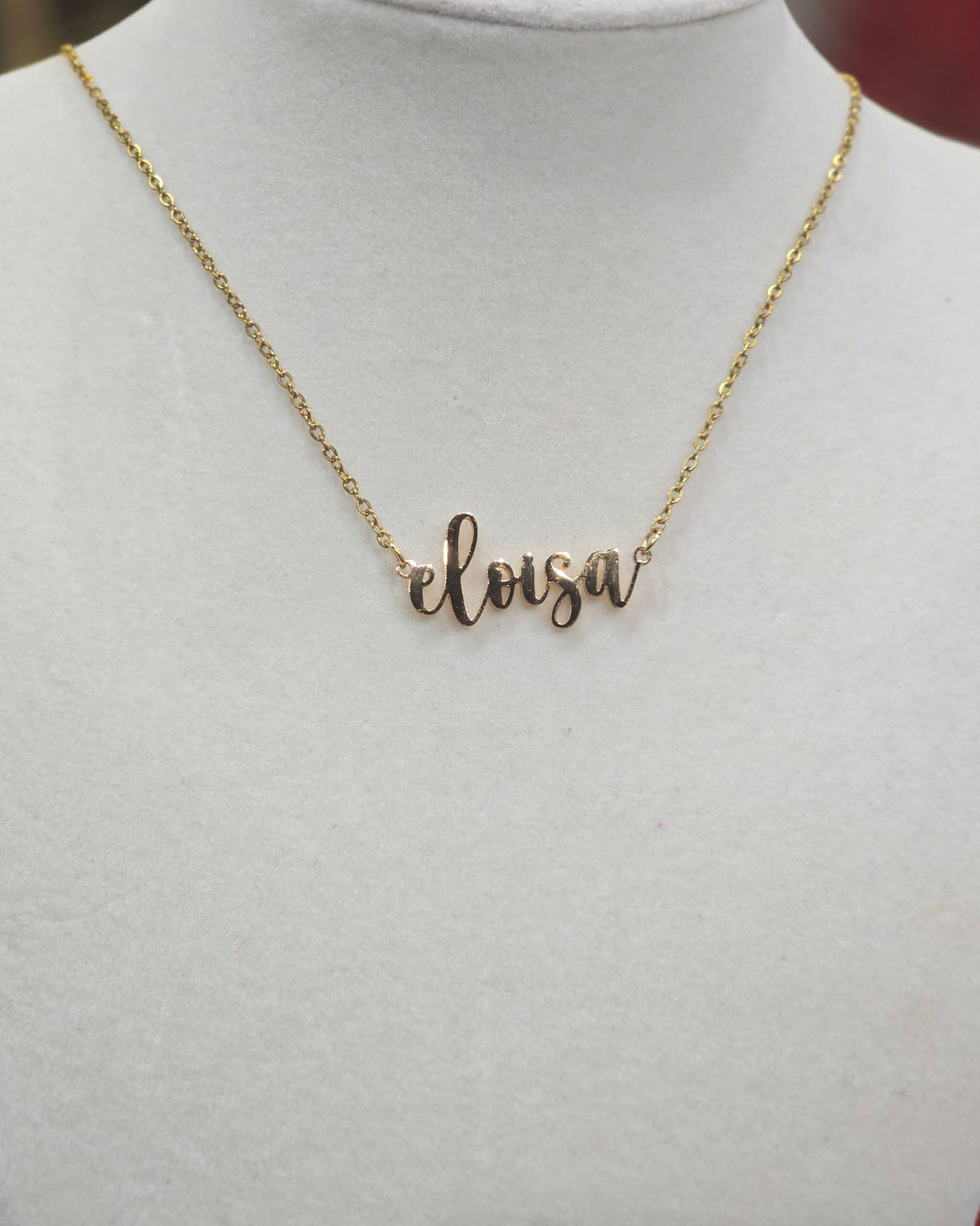 A delicate gold chain necklace with a cursive nameplate spelling 'eloisa', displayed on a white jewelry bust.